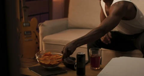 Smiling guy relaxes on couch in man's living room, reaches for bowl of chips guy 動画素材 200791321