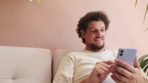 Smiling guy sitting on the sofa in cozy living room and using the phone Stock Footage 252030964