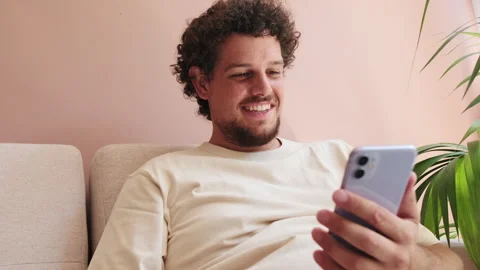 Smiling guy sitting on the sofa in cozy living room scrolls across the phone Stock Footage 252207382