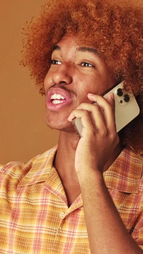 Smiling guy speaks emotionally on mobile phone, isolated on brown background in Stock Footage 253933956