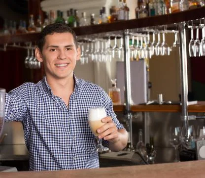 Smiling handsome bartender standing behind the bar counter and h Stock Photos