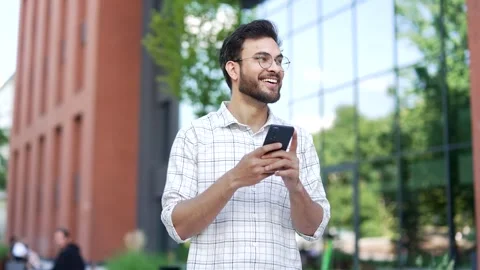 Smiling handsome man is using a mobile phone while walking on the street near  Video stock 306309398