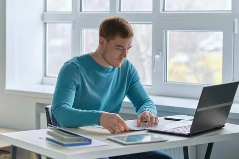Smiling handsome young guy sitting at table, looking at computer screen. Focused Stock Photos