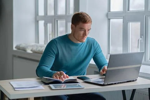 Smiling handsome young guy sitting at table, looking at computer screen. Focused Stock Photos