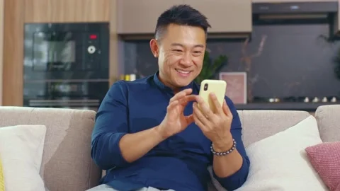 Smiling happy young asian man use phone sitting on sofa at home. Smartphone Stock Footage 141007259