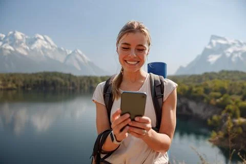 A smiling hiker uses a smartphone while enjoying the beautiful lake and Stock Photos