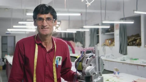 Smiling Indian tailor man facing the camera in a garment workshop Stock Footage 243633136