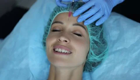 Smiling lady gets injection of botox in her forehead, cosmetologist using Stock Photos
