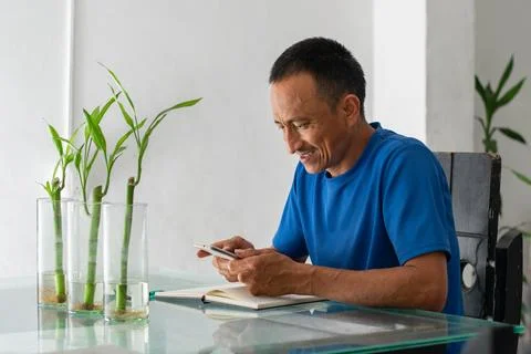 Smiling Latino man at a table using a smartphone, informal home scene, with.. Stock-Fotos