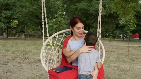 A smiling little boy embraces his mother, who is sitting on a swing in the park  Video stock 115716620