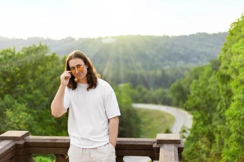 Smiling long-haired man in a white tshirt adjusts sunglasses on a balcony. .. 스톡 사진