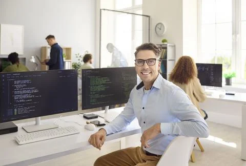 Smiling male developer at workstation with code on monitors in modern office Photos