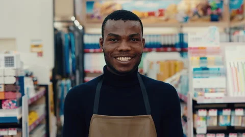 Smiling male store clerk wearing an apron, standing in a well-organized retail Stock-Footage 313359953