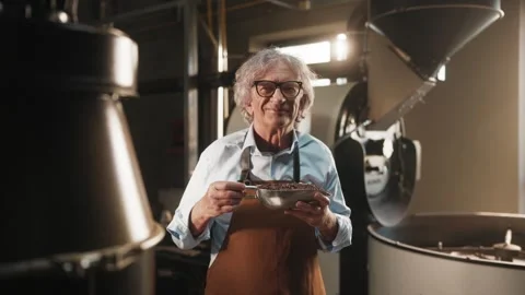 Smiling man in brown apron and blue shirt holding metal bowl of roasted coffee Stock Footage 315072326