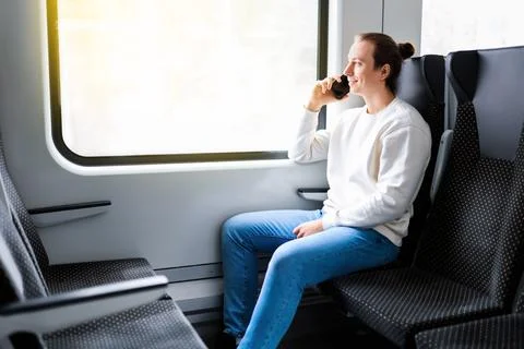 Smiling man with a bun talking on a smartphone while sitting by the window .. 스톡 사진