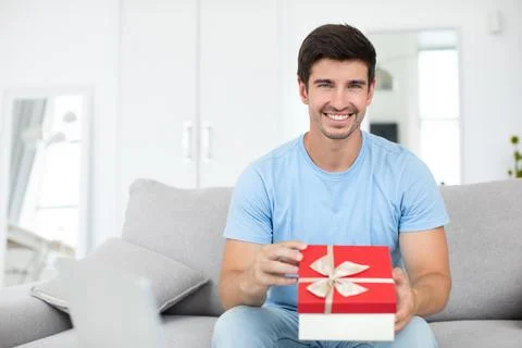 Smiling man with a gift in his hands on the couch at home. Stock Photos