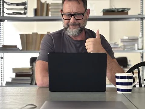 Smiling man giving thumbs up while working on laptop at casual home office de Foto stock