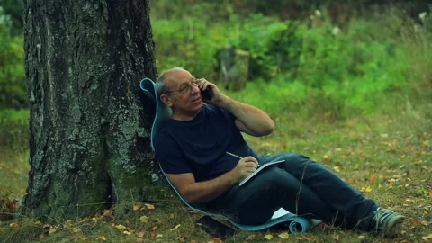 A smiling man in glasses sits under a tree in the park and writes in a notebo Vidéo 97528039