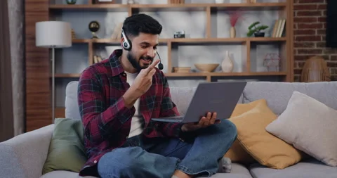 Smiling man in headphones using laptop sitting on sofa at home. Hispanic male in Stock Footage 225543723