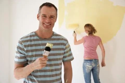 Smiling Man Holding Paintbrush While Woman Paints Wall With Roller Stock Photos