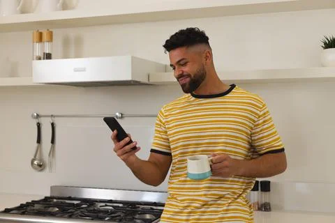 Smiling man in kitchen holding coffee mug and checking smartphone 写真素材