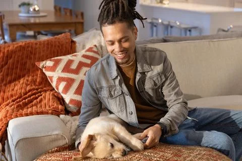 Smiling man leaning forward on sofa at home, wearing denim jacket petting small Stock Photos