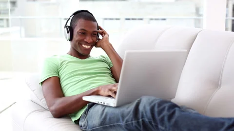 Smiling man lying on couch listening to music using laptop Stock Footage