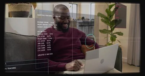 Smiling man in maroon sweater checking metrics on laptop at home on gray sofa, 스톡 사진