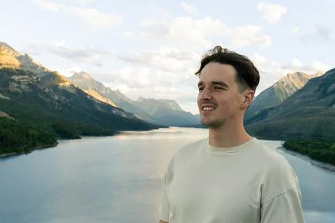 A smiling man in a neutral tee enjoys a lakeside mountain vista at twilight. Stock Photos