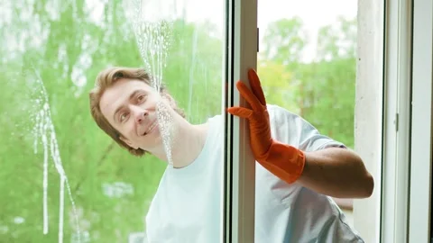 Smiling man in orange gloves uses a squeegee to wash a window pane.  Video stock 335747007