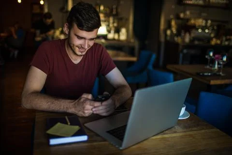 Smiling man owner using messenger on mobile phone while sitting with laptop c Stock Photos