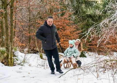 Smiling man pulling sled with his wife and daughter through snow in winter fo Stock Photos