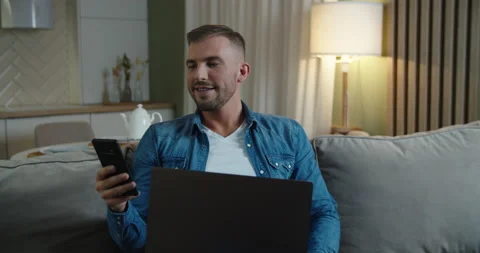 Smiling man reads a message on a mobile phone, in his hands a laptop. Kitchen Stock-Footage 147221109