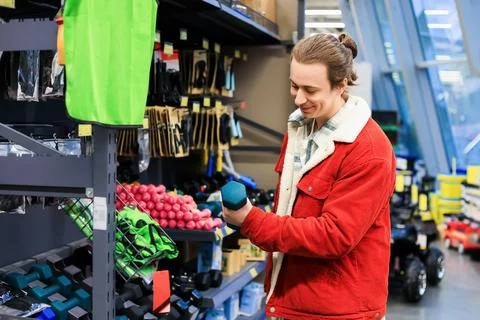 Smiling man in red jacket holding blue dumbbell while shopping for sports e.. Stock Photos