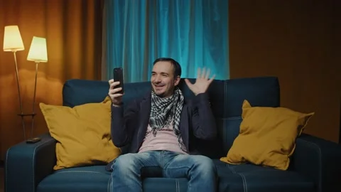 Smiling man sits on sofa at home and answers video call on smartphone, share Stock Footage 171925855
