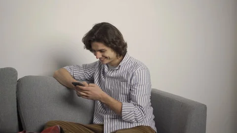 Smiling man sitting on couch and using smartphone in living room Stock Footage 124525164