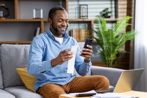 Smiling man sitting on couch using smartphone and holding paper, enjoying online 写真素材