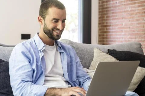Smiling man sitting on a couch using a laptop at home, working remotely and.. Foto stock