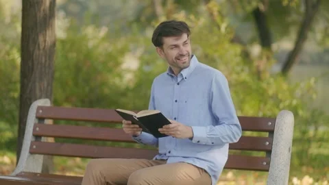 Smiling man sitting on park bench reading a book in autumn sunlight. 스톡 동영상 332299112