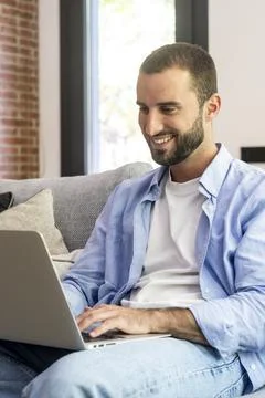 Smiling man on a sofa using a laptop for remote work and online shopping, c.. Stock Photos