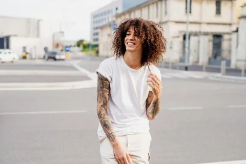 Smiling man with tattooed arms and long brown curly hair standing on a street. Stock Photos