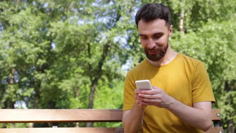 Smiling man texting on smartphone on bench in park 库存影片 136114352