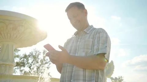 Smiling man use of mobile phone at part near the fountain at sunlight cellphone Stock Footage 116997294
