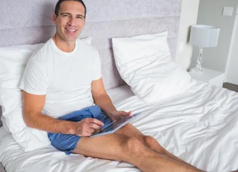 Smiling man using his tablet pc sitting on bed 스톡 사진
