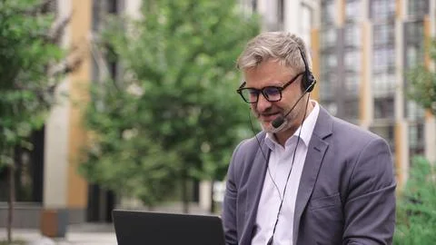 Smiling man using laptop and headset, talking online outdoors. Slow motion Foto stock