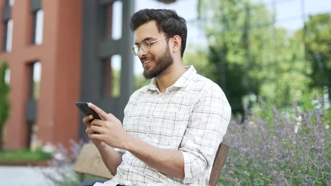 Smiling man is using a mobile phone while sitting on a bench on street near Stock Footage 301118059