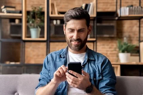 Smiling man using mobile phone scrolling network feed sitting on sofa at home Stock Photos
