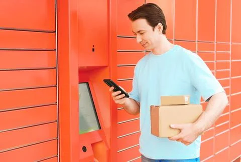 Smiling man using a smartphone at an automated orange parcel locker to pick.. Stock Photos