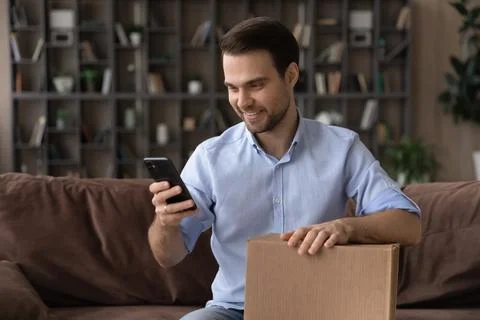 Smiling man using smartphone, holding cardboard box, unpacking parcel 写真素材