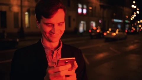 Smiling man using smartphone at night in city. Face illuminated by red light Stock Footage 80009381